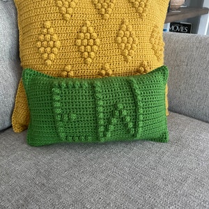 May include: Two crocheted throw cushions, one yellow with a textured pattern and one green with a textured pattern. The cushions are on a grey sofa.
