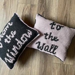 May include: Two crocheted cushions with black text on a light pink and black background. The text on the cushions reads "Open the window" and "To the wall".