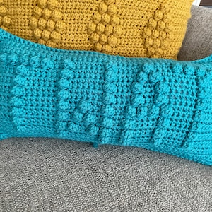 May include: Two crocheted throw pillows, one teal blue and one mustard yellow, with a textured, bumpy surface. The pillows are on a gray couch.