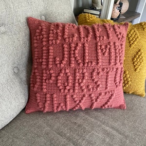 May include: Two crocheted throw pillows, one in a deep rose color and the other in a mustard yellow. Both pillows have a textured, bumpy surface.
