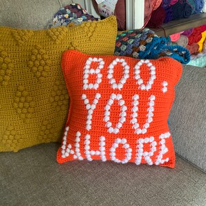 May include: Orange crocheted pillow with white crocheted letters spelling "BOO YOU WHERE" on a gray couch.