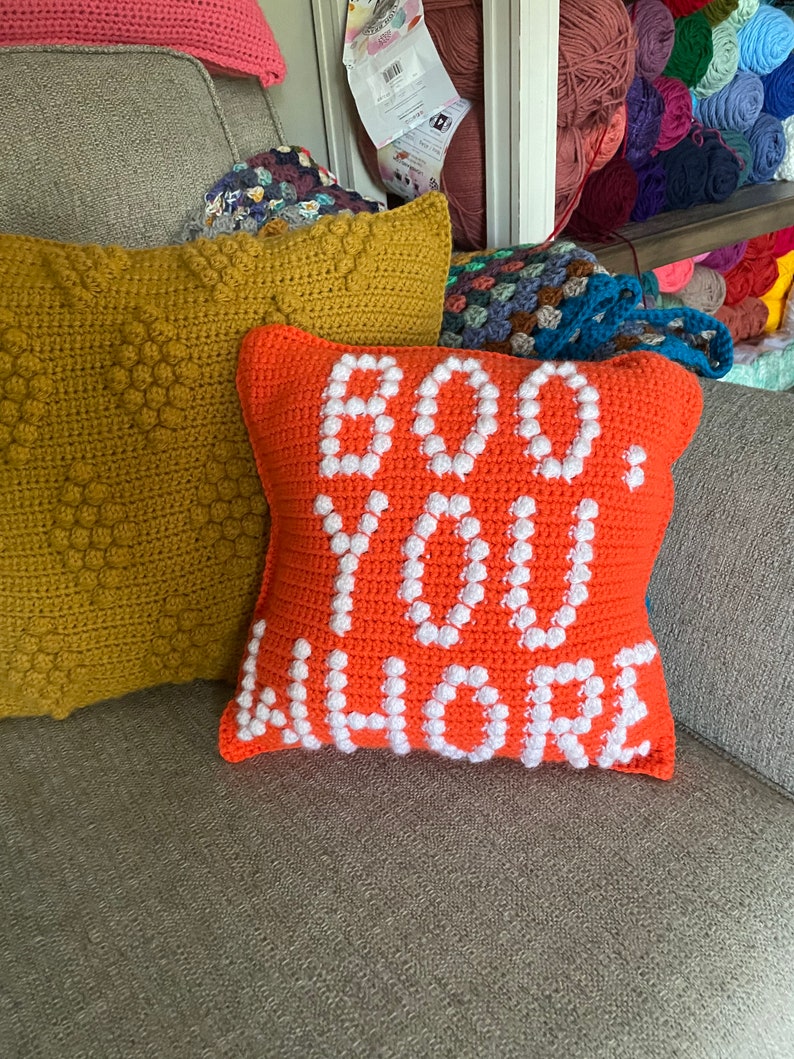 May include: Orange crocheted pillow with white crocheted letters spelling "BOO YOU WHERE" on a gray couch.