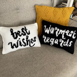 May include: Three crocheted throw pillows with different designs. The first pillow is white with black text that says "best wishes". The second pillow is black with white text that says "warmest regards". The third pillow is yellow with a textured pattern.