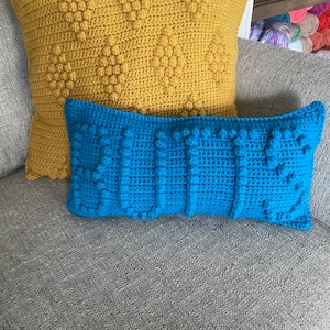 May include: Two crocheted throw pillows, one yellow and one blue, with a textured, bumpy surface. The pillows are on a gray couch.