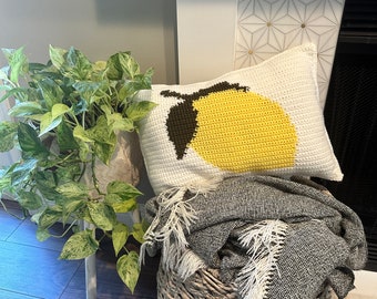 May include: A crocheted lemon pillow with a white background and yellow lemon design. The pillow is sitting on a woven basket with a gray and white blanket.