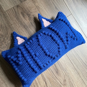 Puede incluir: Una almohada de crochet azul con dos orejas de gato de fieltro rosa. La almohada es texturizada y tiene una superficie ligeramente abultada.