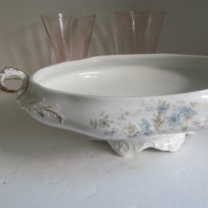 May include: White oval serving bowl with blue floral pattern and gold trim. The bowl has two handles and sits on a decorative base. Two pink glasses are in the background.