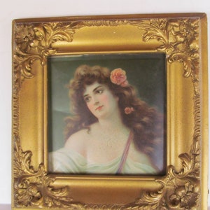 May include: A framed portrait of a woman with long, flowing auburn hair adorned with pink roses. The portrait is set within an ornate, square gold-colored frame with detailed corner embellishments. The woman is wearing a light-colored gown.