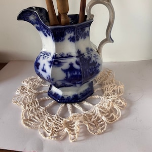 Op de afbeelding: Een blauwe en witte porseleinen kan met een bloemenmotief, met daarin een verzameling blauwe en witte keramische keukenspullen met houten handvatten. De kan staat op een wit gehaakt onderzetter.