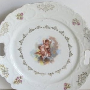 May include: White porcelain serving plate with gold trim and a cherub design. The plate has two handles.