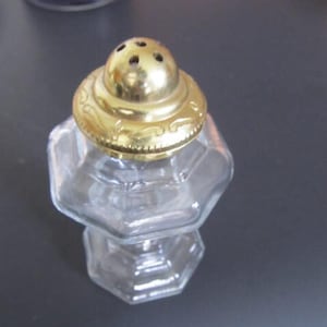 Puede incluir: Un salero de vidrio transparente con tapa de metal dorado y diseño decorativo. La tapa tiene cinco agujeros para dispensar sal.