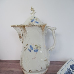 May include: A white porcelain teapot with a gold trim and blue floral design. The teapot has a lid with a finial and a handle on the side. The teapot is sitting on a wooden surface.
