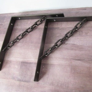 May include: Two black metal shelf brackets with a chain design. The brackets are attached to a wooden surface.