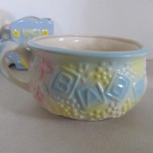 May include: A pastel-colored ceramic baby cup with a handle, featuring the word "BABY" in raised letters. The cup has a light blue rim and accents, with embossed floral and dot patterns. A small ceramic lamb and a blue carriage are in the background.