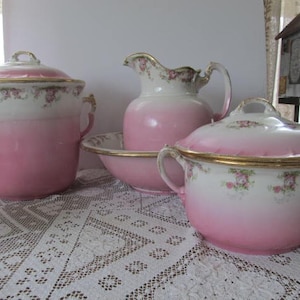 May include: A set of vintage pink and white porcelain bathroom accessories with gold trim. The set includes a covered chamber pot, a pitcher, and a wash basin.
