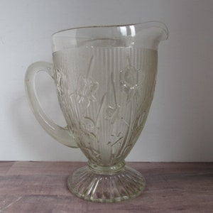 May include: A clear glass pitcher with a textured floral design. The pitcher has a wide base and a handle.