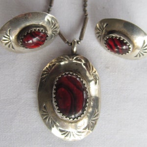 May include: A silver necklace and earrings set with red stone accents. The necklace features a pendant with an oval-shaped stone surrounded by a silver frame. The earrings are small and round with a red stone in the center.