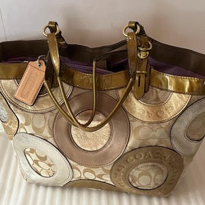 May include: A large, brown and gold Coach tote bag with a circular pattern. The bag has a top zipper closure and two top handles.