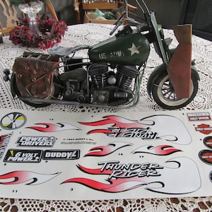 May include: A green toy motorcycle with a sidecar, a US Army star, and the words "Thunder Rider" on the side. The motorcycle is on a white sheet with a sheet of stickers for the motorcycle. The stickers include the words "Power Drivers", "6 Volt Power", "Buddy L", and "Harley Davidson".