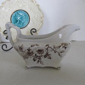 May include: White porcelain gravy boat with a brown floral design. The boat has a curved handle and a spout.