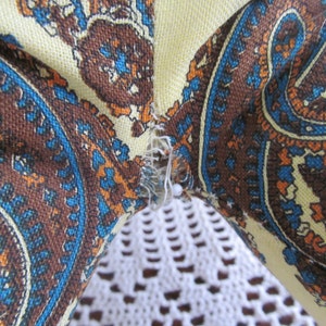 K&ouml;nnte beinhalten: Nahaufnahme eines Stoffes mit Paisley-Muster in Braun-, Blau- und Oranget&ouml;nen. Der Stoff ist mit einer wei&szlig;en Spitzenborte zusammengen&auml;ht.