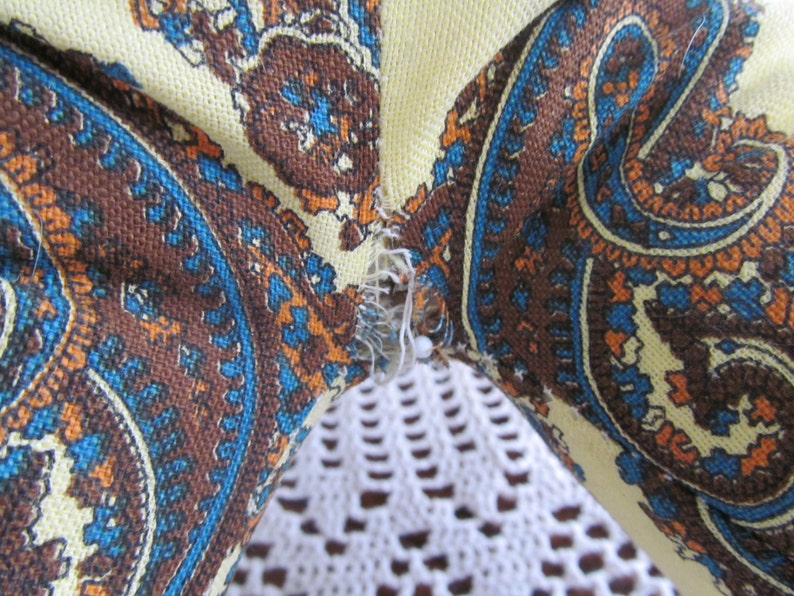 K&ouml;nnte beinhalten: Nahaufnahme eines Stoffes mit Paisley-Muster in Braun-, Blau- und Oranget&ouml;nen. Der Stoff ist mit einer wei&szlig;en Spitzenborte zusammengen&auml;ht.