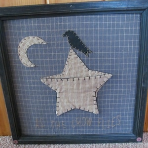 May include: Framed wall art featuring a blue grid background with a beige star, crescent moon, and a black crow. The phrase "AS THE CROW FLIES" is stitched at the bottom. The frame is dark green with pink button accents.