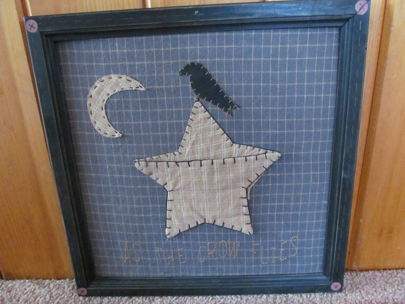 May include: Framed wall art featuring a blue grid background with a beige star, crescent moon, and a black crow. The phrase "AS THE CROW FLIES" is stitched at the bottom. The frame is dark green with pink button accents.