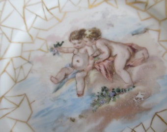 Peut inclure: Une assiette en porcelaine vintage avec une délicate bordure dorée et une scène peinte de deux chérubins jouant dans un paysage bleu et blanc.