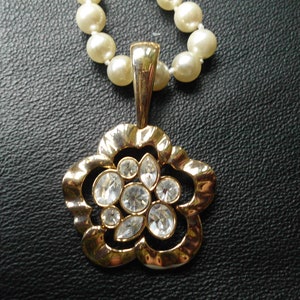 Pu&ograve; includere: Un ciondolo dorato a forma di fiore con strass trasparenti pende da una collana di perle. La collana e il ciondolo sono su una superficie nera.