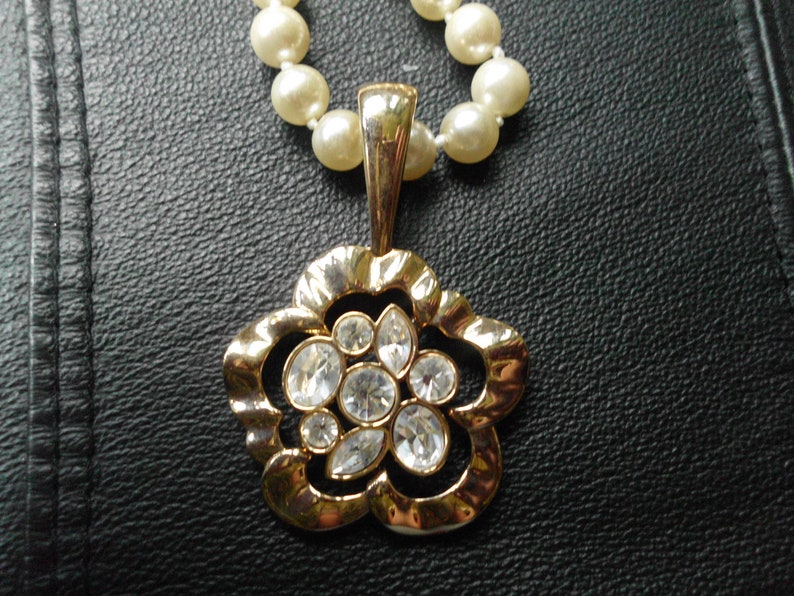 Pu&ograve; includere: Un ciondolo dorato a forma di fiore con strass trasparenti pende da una collana di perle. La collana e il ciondolo sono su una superficie nera.