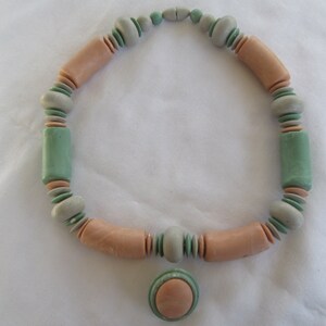 May include: A necklace made of pastel pink, green, and gray ceramic beads. The necklace has a pendant in the shape of a large, flat, pastel pink circle.