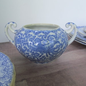 May include: A blue and white porcelain sugar bowl with two curved handles. The bowl features a detailed pattern of swirling designs and a bird motif. Several matching plates and a covered dish are visible, suggesting a tea set.