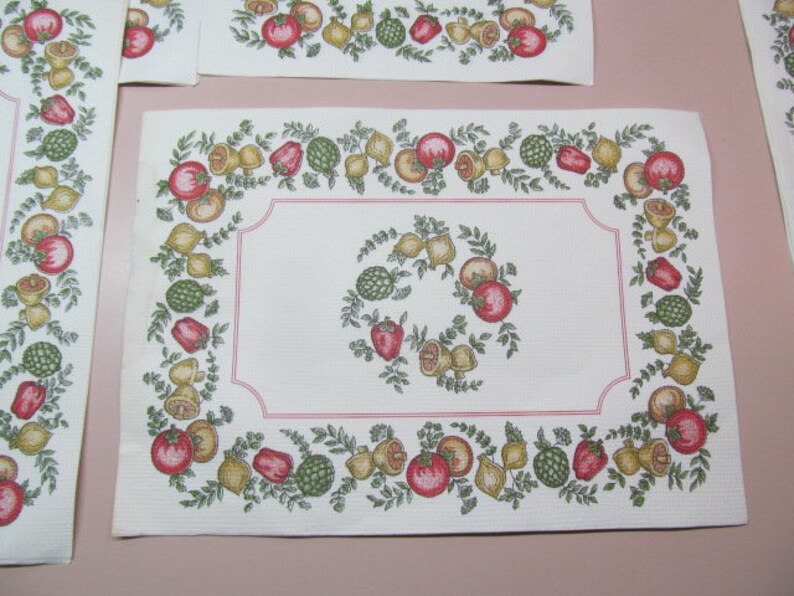 May include: White placemat with a red, green, and yellow border featuring a repeating pattern of fruit and vegetables. The border is outlined in pink.