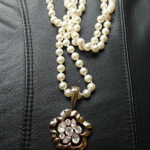 Pu&ograve; includere: Una collana di perle bianche con un ciondolo a forma di fiore dorato con strass trasparenti. La collana &egrave; su uno sfondo di pelle nera.