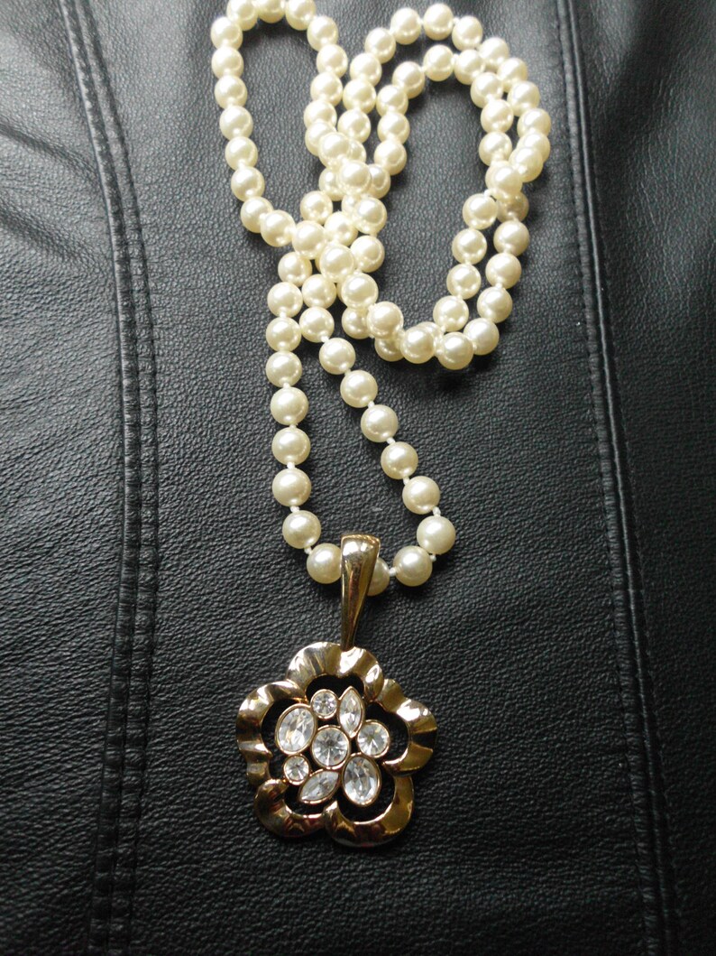 Pu&ograve; includere: Una collana di perle bianche con un ciondolo a forma di fiore dorato con strass trasparenti. La collana &egrave; su uno sfondo di pelle nera.
