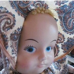 K&ouml;nnte beinhalten: Eine Porzellanpuppe mit blauen Augen und blonden Haaren, die einen wei&szlig;en und braunen Paisley-Muster-Hut tr&auml;gt.