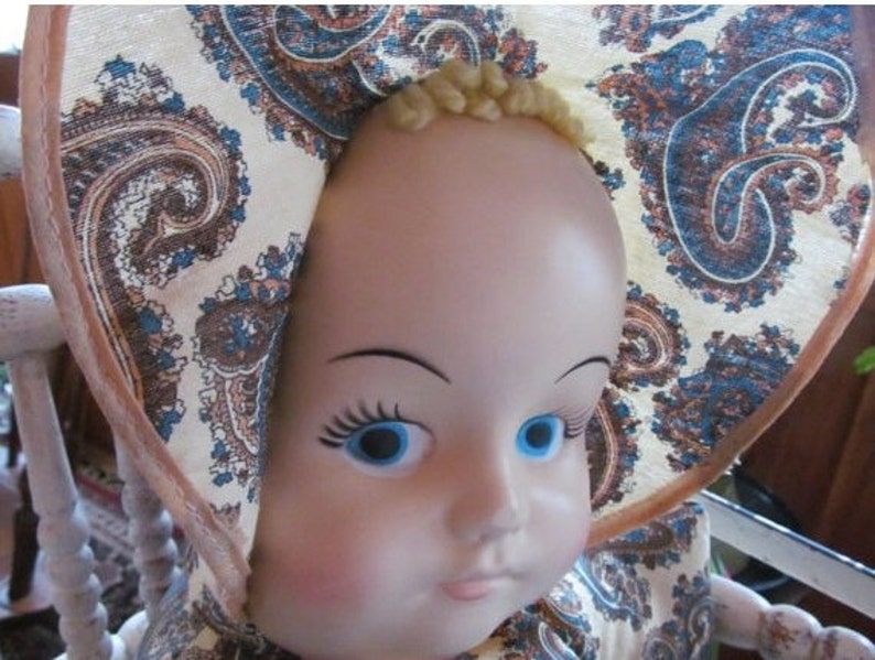 K&ouml;nnte beinhalten: Eine Porzellanpuppe mit blauen Augen und blonden Haaren, die einen wei&szlig;en und braunen Paisley-Muster-Hut tr&auml;gt.