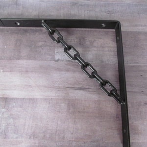 May include: Black metal shelf bracket with a black chain attached to it. The bracket is mounted to a light wood surface.