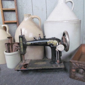 Op de afbeelding: Een vintage zwarte naaimachine met gouden versiering en een witte keramische pot met roestige spijkers. De naaimachine staat op een houten oppervlak naast een witte keramische kan en een witte keramische kruik.