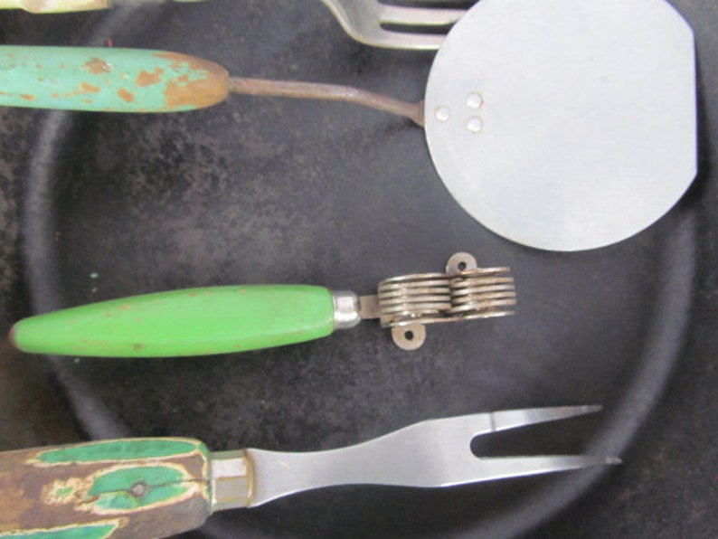 Puede incluir: Un juego de cuatro utensilios de cocina vintage con mangos verdes y blancos. Los utensilios incluyen una esp&aacute;tula, un tenedor, un cuchillo y un machacador de patatas.