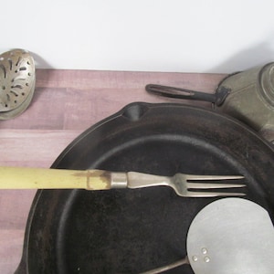 May include: A cast iron skillet with a wooden handle, a green and white handled fork, a silver slotted spoon, and a silver spatula. The skillet is on a wooden surface.