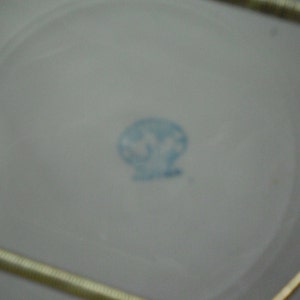 May include: A close-up of the bottom of a white ceramic plate with a blue circular mark in the center. The mark contains a stylized design of a bird with a crown above it. The text "Made in Japan" is visible below the design.