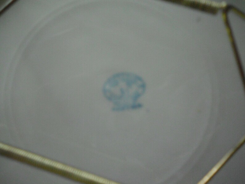 May include: A close-up of the bottom of a white ceramic plate with a blue circular mark in the center. The mark contains a stylized design of a bird with a crown above it. The text "Made in Japan" is visible below the design.