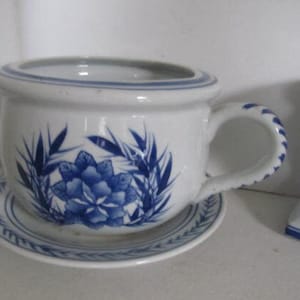 May include: A white ceramic planter with a blue and white floral design. The planter has a wide, round base and a handle on one side. It is sitting on a matching saucer.