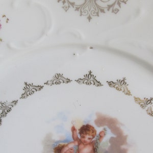 May include: A white porcelain plate with a gold trim and a floral design. The center of the plate features a painting of a cherub.