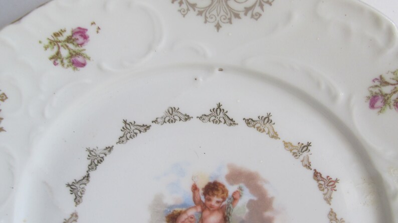 May include: A white porcelain plate with a gold trim and a floral design. The center of the plate features a painting of a cherub.