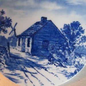 May include: Blue and white porcelain plate with a hand-painted design of a small cottage with a windmill in the background. The cottage has a blue roof and white trim. The windmill is also blue and white. The plate is surrounded by a blue and white border.