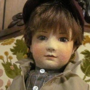 May include: A vintage porcelain doll with brown hair wearing a brown felt hat and a tan jacket with a plaid shirt underneath. The doll has blue eyes and rosy cheeks.