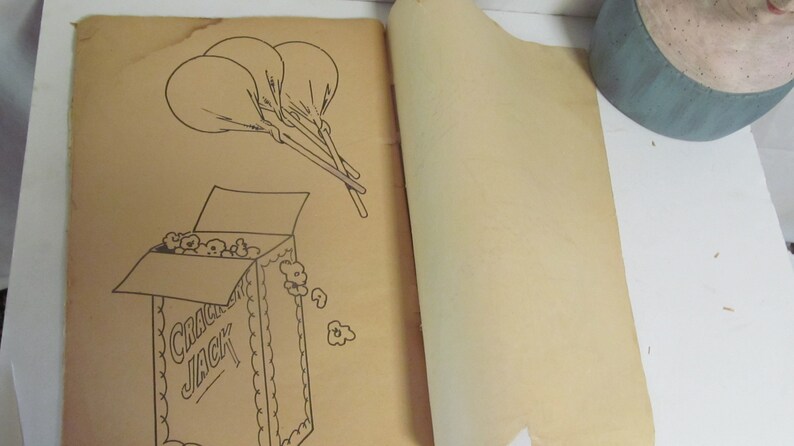 Puede incluir: Una p&aacute;gina de libro para colorear vintage con una ilustraci&oacute;n en blanco y negro de una caja de palomitas de ma&iacute;z Cracker Jack con la tapa abierta y las palomitas de ma&iacute;z derram&aacute;ndose. Hay tres globos atados juntos con cuerdas sobre la caja.